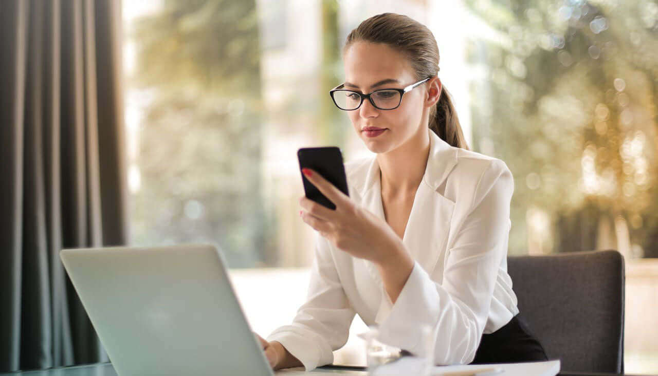 Smart Security for Moms: Keep Your Home Safe Anywhere | corporate woman wearing glasses in front of laptop checking her smart phone