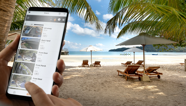hands holding smartphone showing real time footage of  Toucan floodlight camera while person is on vacation at the beach