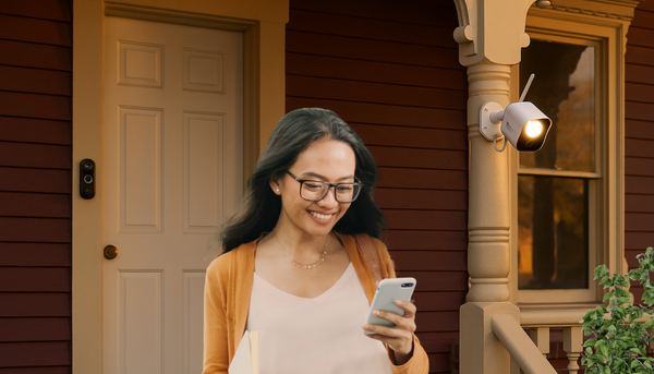 woman looking at her phone and walking down the front steps of the burgundy victorian house with the doorbell v3 near the white front door and the floodlight camera by the porch near a plant