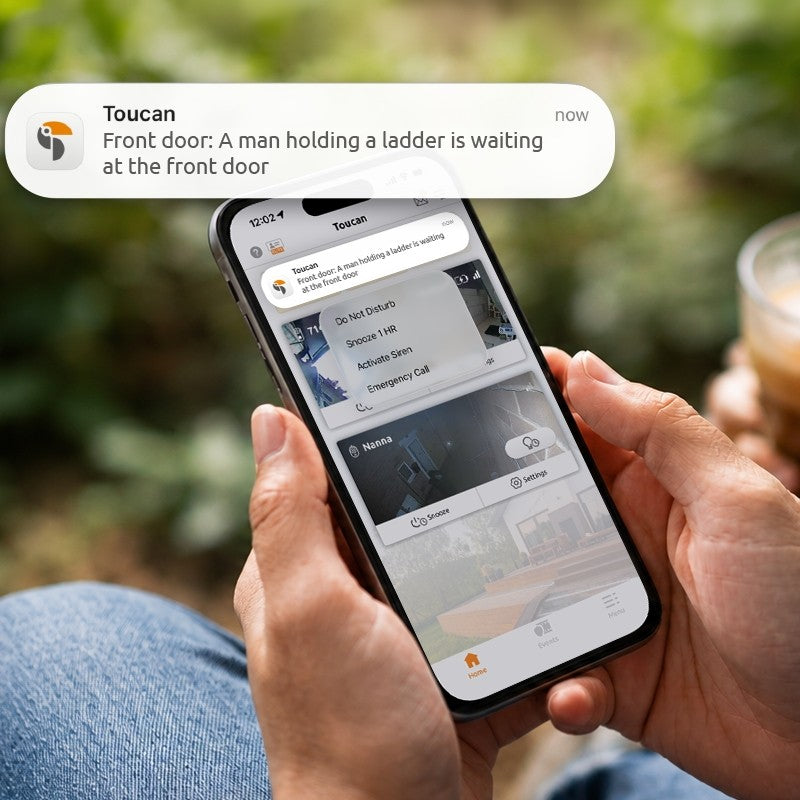 A person holds a smartphone showing a security app notification overlay from Toucan, with a blurred outdoor background and part of a drink visible.